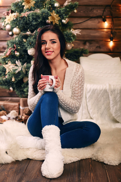 Beautiful young woman drinking tea at the Christmas tree. Beautiful girl celebrates Christmas with a cup of cocoa in front of tree over living room - Фото, зображення