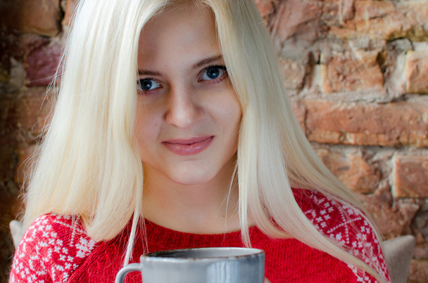 Блондинка в красном свитере возле кирпичной стены с чашкой в руках
 - Фото, изображение
