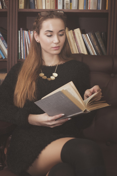 young beautiful girl in a black dress sitting on a leather sofa in the library with books - Foto, Bild