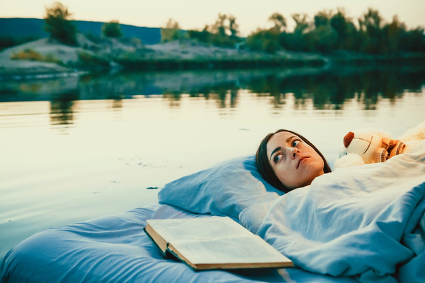 Pretty woman is laying on the floating bed with a book and toy.  - Фото, зображення