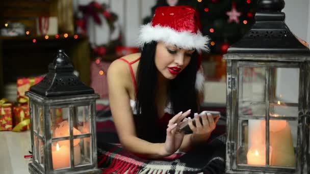 Ευτυχισμένος ελκυστική χαμογελαστό νεαρή γυναίκα με το κινητό στο πάτωμα στο δωμάτιό της ζωής μπροστά από ένα χριστουγεννιάτικο δέντρο - Πλάνα, βίντεο