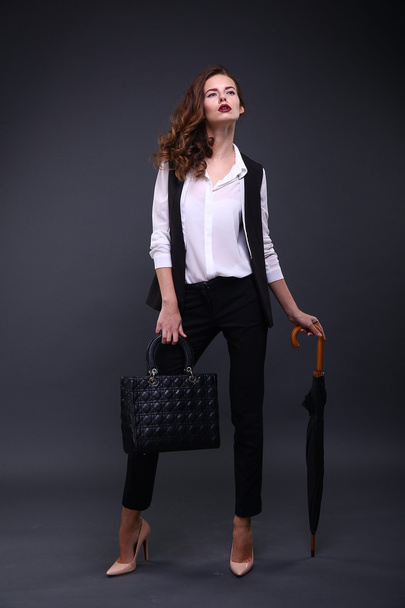 Pretty female business lady in a black jacket white shirt, umbrella, handbag on a dark background. Studio shot. - Foto, afbeelding