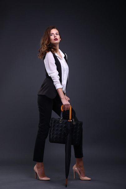 Pretty female business lady in a black jacket white shirt, umbrella, handbag on a dark background. Studio shot. - Fotó, kép