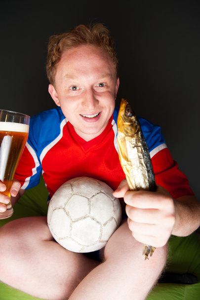 Young man holding soccer ball, beer and stockfish, watching tv translating of game at home wearing sportswear - Photo, Image