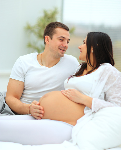 man looks lovingly at his wife and strokes her baby bump, lying on her white bed - Photo, image
