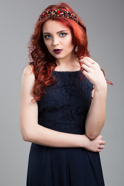 Beautiful red-haired fashion model posing in evening dress and in the diadem over dark background. female gestures of seduction. body language. touching her hair. Wavy Red Hair. Fashion Girl Portrait. - Foto, Imagen