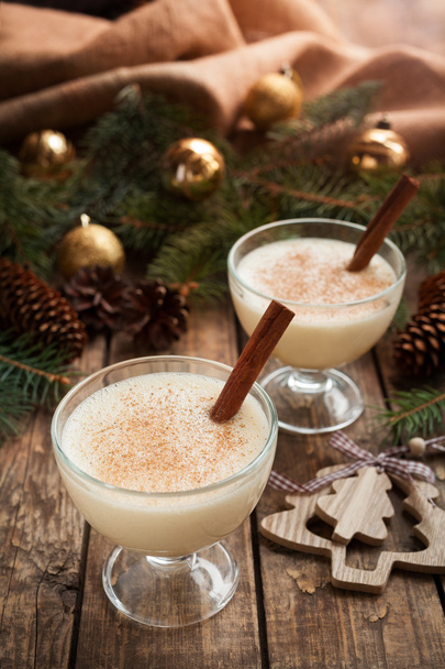 Eggnog traditional homemade egg, milk, rum, vanilla alcohol liqueur preparation recipe in two glass cups with cinnamon sticks on wooden vintage table. Shallow depth of field. - Fotó, kép