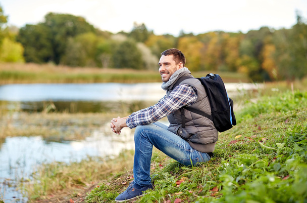 smiling man with backpack resting on river bank - Фото, изображение