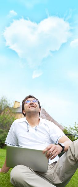 Un uomo sorridente con il computer portatile all'aperto seduto nel suo cortile e guardando verso la nuvola di forma di cuore sopra la testa
 - Foto, immagini