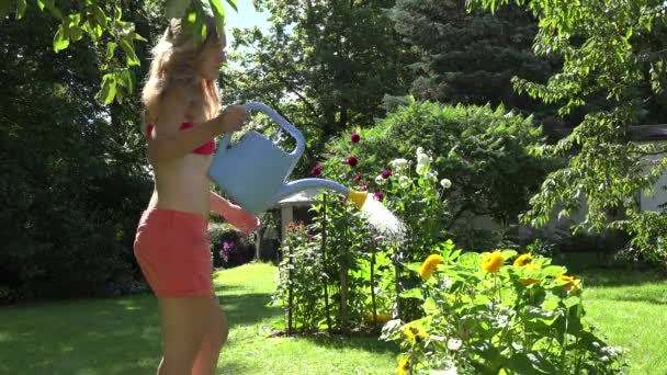 σέξι γυναίκα κηπουρός σε σορτς και σουτιέν με νερό μπορεί να ποτίζει ανθίζουν ηλιοτρόπια στην αυλή. 4K - Πλάνα, βίντεο