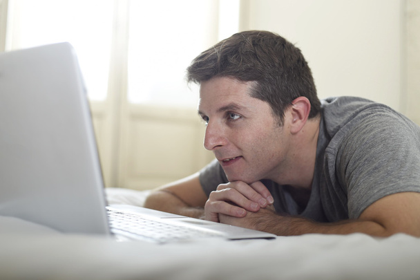 portrait of young attractive man lying on bed watching on line movie in computer laptop at home in internet concept - Photo, Image