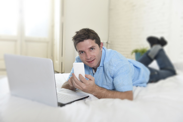 young attractive man lying on bed using mobile phone and computer laptop working from home - Foto, Imagen