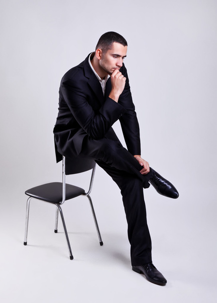灰色の背景上の椅子に座ってエレガントな若いビジネスマン - 写真・画像