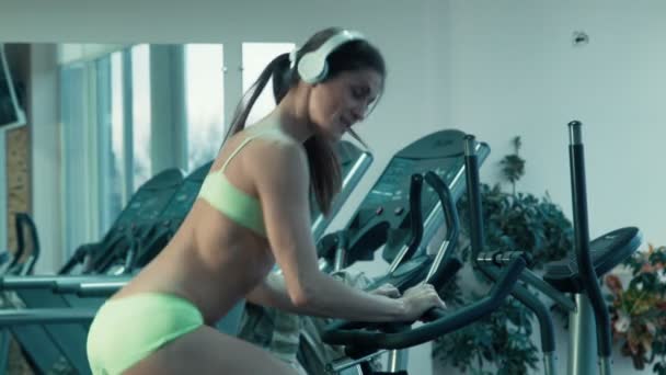 Αθλητική θηλυκό με ακουστικά pedaling σε ο προσομοιωτής σε στατικό ποδήλατο στο γυμναστήριο. - Πλάνα, βίντεο