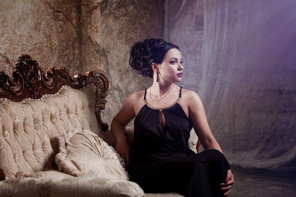 attractive young brunette in stylish black evening dress, is back  half a turn, sitting on the sofa - 写真・画像