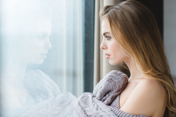 Beautiful woman in grey knitted coverlet looking out of window - Foto, afbeelding