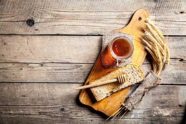 Honey in a jar, slice of bread, wheat on an old vintage planked wood table. - Fotoğraf, Görsel