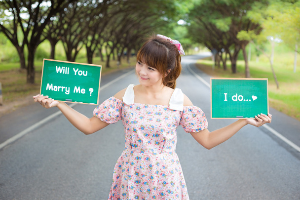 Cute woman hand holding green board sign with text " will you marry me" and "i do"on road and tree, Smiling female model. - Photo, Image
