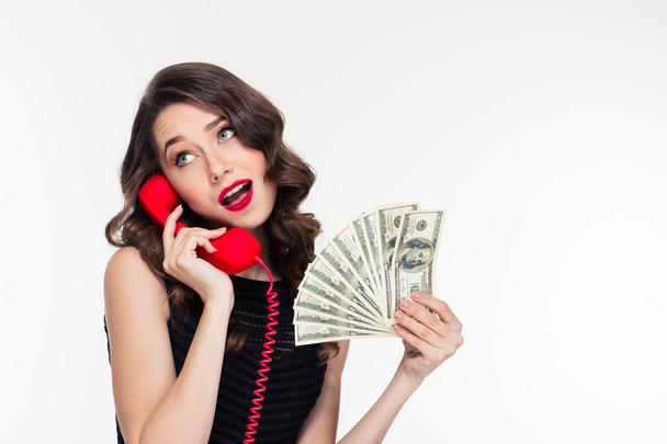 Attractive joyful curly female holding money and talking on telephone - Фото, изображение