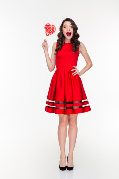 Cheerful happy woman in red dress holding heart shaped lollipop - Fotó, kép