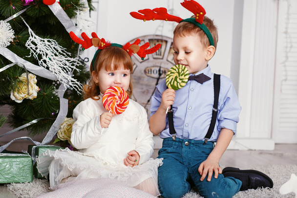 Девочка и мальчик в оленьих рогах едят леденцы
.  - Фото, изображение