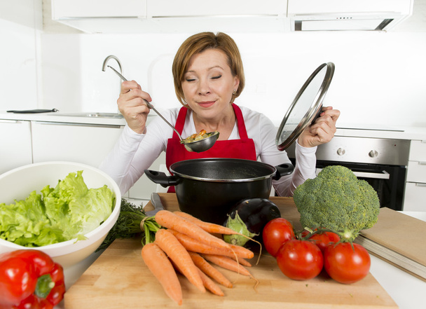 молодая счастливая домохозяйка в красном фартуке на домашней кухне держит кастрюлю дегустируя горячий суп
 - Фото, изображение