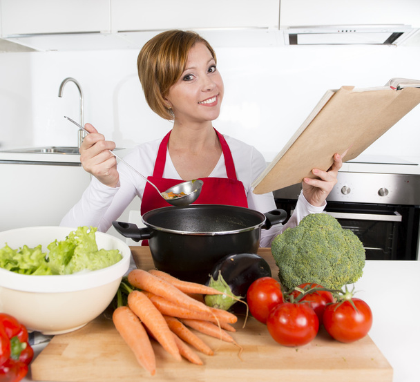 attraktive Köchin bereitet Gemüseeintopfsuppe vor und liest Rezeptkochbuch in der heimischen Küche  - Foto, Bild