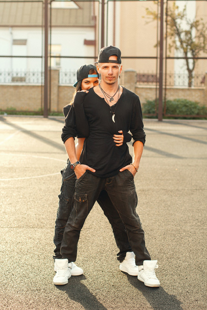 Mujer bailarina de hip-hop en vestido negro con estilo con cadena "Loves" y una gorra. Hombre de pie en una pose de baile en el fondo
. - Foto, imagen