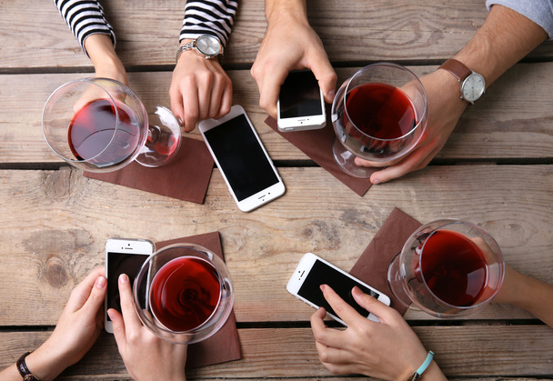 Four hands with smart phones holding glasses with red wine, on wooden table background - Фото, зображення
