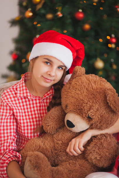 holidays, presents, christmas, childhood and people concept - smiling girl in santa helper hat with teddy bear over lights background - Foto, Imagem