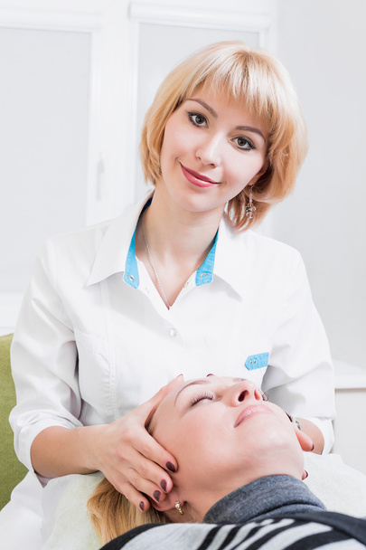  mujer rubia esteticista permanente en una bata blanca en la oficina con patsientom.delaet masaje facial. ¿Doctor? sonriendo
 - Foto, Imagen