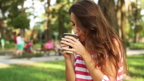 hermosa mujer sentada en un banco en el parque y comiendo un panecillo y un café con leche
 - Imágenes, Vídeo