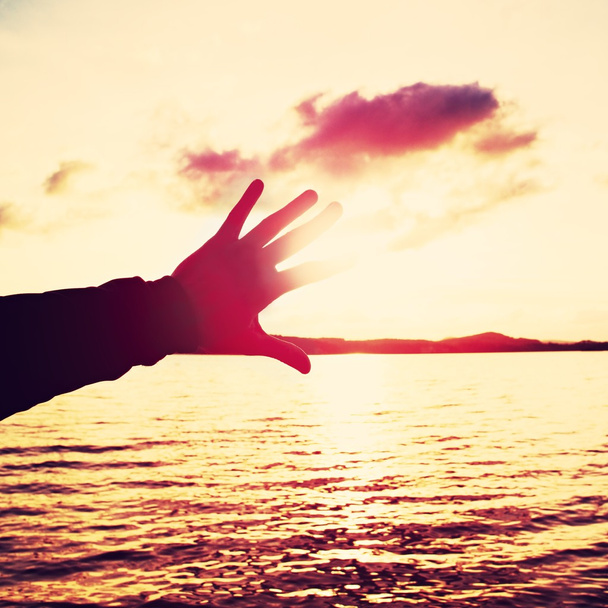 Kéz- és lomg ujjak sziluettje próbálja megérinteni a nap óceán feletti naplementében. Élénk színek. - Fotó, kép