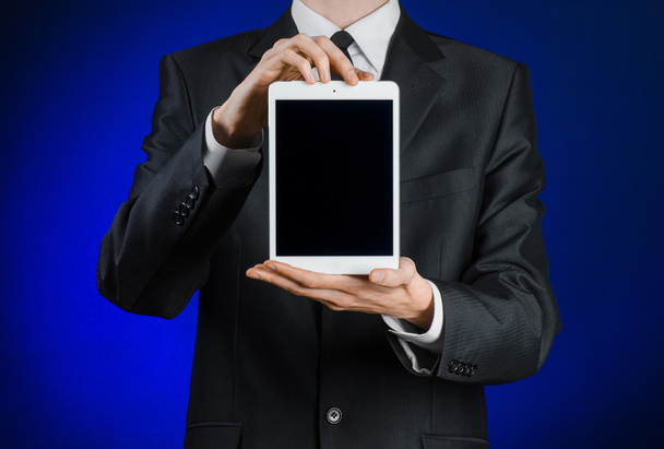 Presentation and business theme: a man in a black suit holding a white tablet touch computer gadget with touch blank black screen on a dark blue background in studio isolated - 写真・画像
