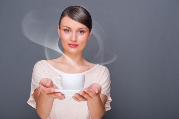 Portrait of young fashionable woman giving hot coffee or tea beverage to camera against grey background - Foto, imagen