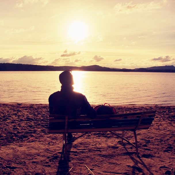 Sam młody człowiek w sylwetka siedzi w słońcu na plaży. Turystyczny odpocząć na ławeczce, drewniane, na jesień jezioro.   - Zdjęcie, obraz