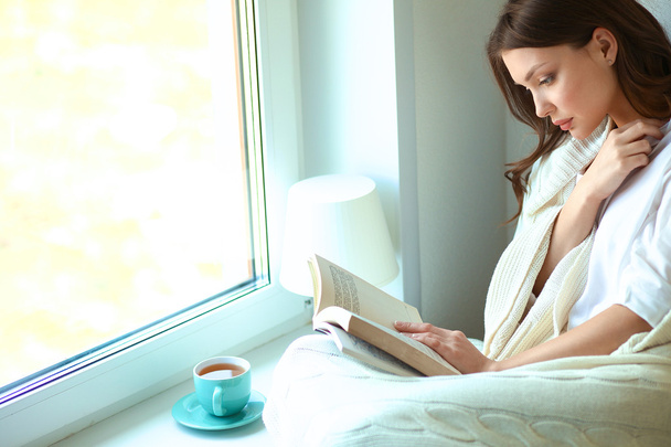 Young woman at home sitting near window relaxing in her living room reading book and drinking coffee or tea - Foto, Imagem