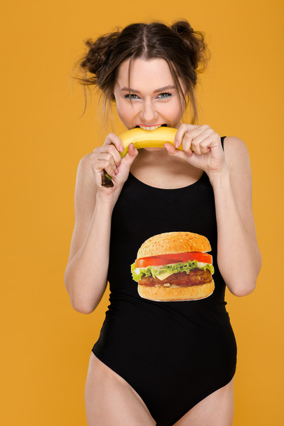 Amusing woman in black designer swimsuit standing and biting banana - Fotografie, Obrázek