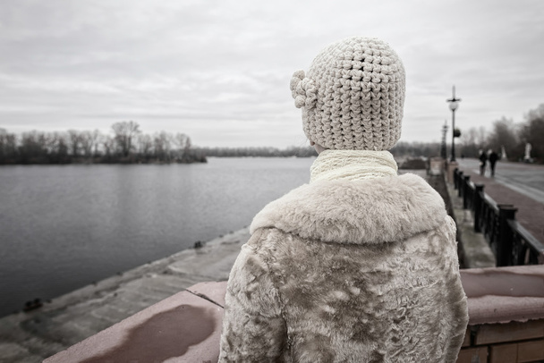 A woman with a wool cap and a fur coat is looking at the river during a cold and sad gray winter morning - Photo, Image