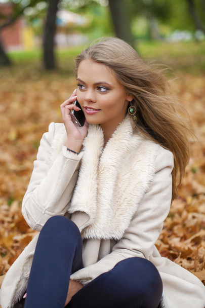 Young happy blonde woman calling by phone in autumn park - Fotó, kép