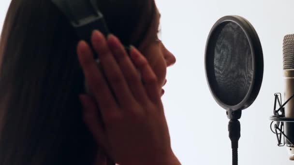 Pretty Girl Singing Into Studio Microphone. The girl is wearing headphones on a white background. In the Low light of the studio. White background. - Footage, Video
