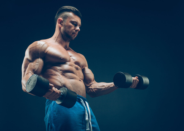 Closeup of a muscular young man lifting dumbbells weights on dark background - Фото, зображення