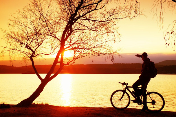 Nuori pyöräilijä siluetti sinisellä taivaalla ja auringonlaskun taustalla rannalla. Kauden päätteeksi järvellä
. - Valokuva, kuva