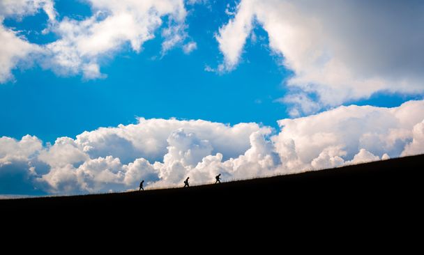 silueta de tres mochileros caminando hacia la cima del fondo nublado de montaña
 - Foto, Imagen