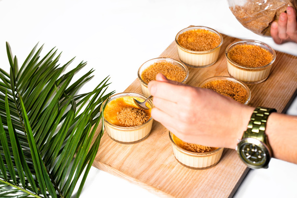 Женские руки делают крем-брюле на деревянном подносе. Традиционный французский ванильный десерт с карамельным сахаром сверху украшенный пальмовыми листьями
. - Фото, изображение