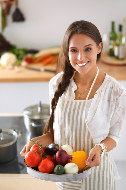 Smiling young woman holding vegetables standing in kitchen - Valokuva, kuva