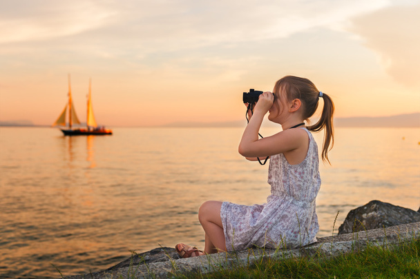 Außenporträt eines netten kleinen Mädchens, das an einem schönen warmen Abend am See spielt und das Boot mit dem Fernglas betrachtet - Foto, Bild
