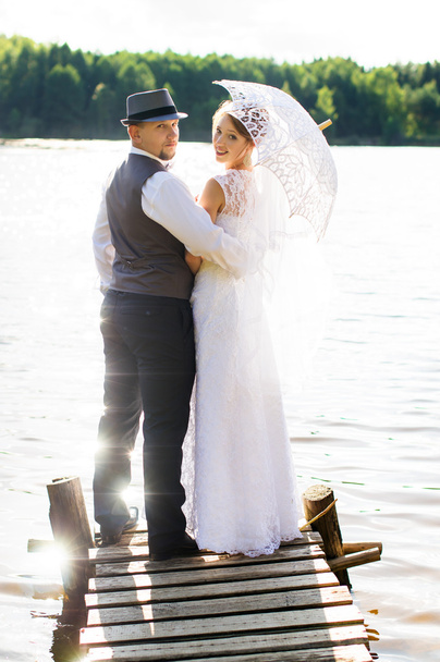 Novia y novio de pie en un puente cerca del agua
 - Foto, Imagen