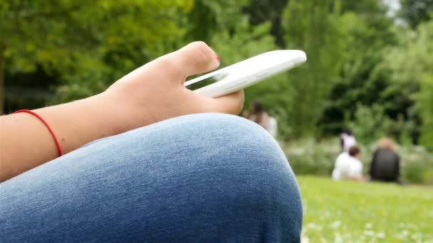 Primer plano de la mujer que utiliza el teléfono móvil sentado en un parque, las manos solamente
 - Metraje, vídeo