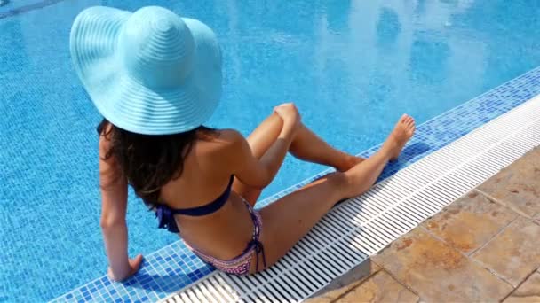 Mladá žena v plavání oblek a modrý klobouk sedí na kraji bazénu a opalovat odběr - Záběry, video
